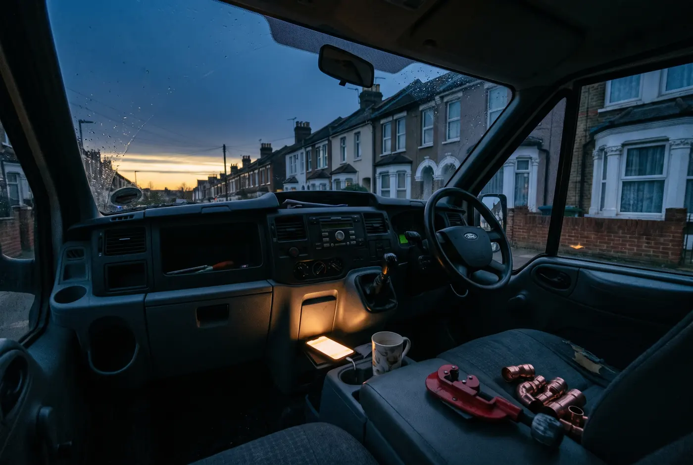 Inside a plumber's van at dawn on a terraced street, with fittings on the passenger seat